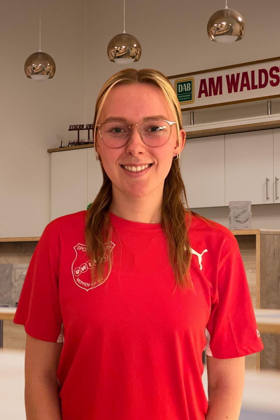 Junge Frau mit Brille und rotem Sportoberteil in einem modernen Raum mit Holzdetails und Deckenleuchten.