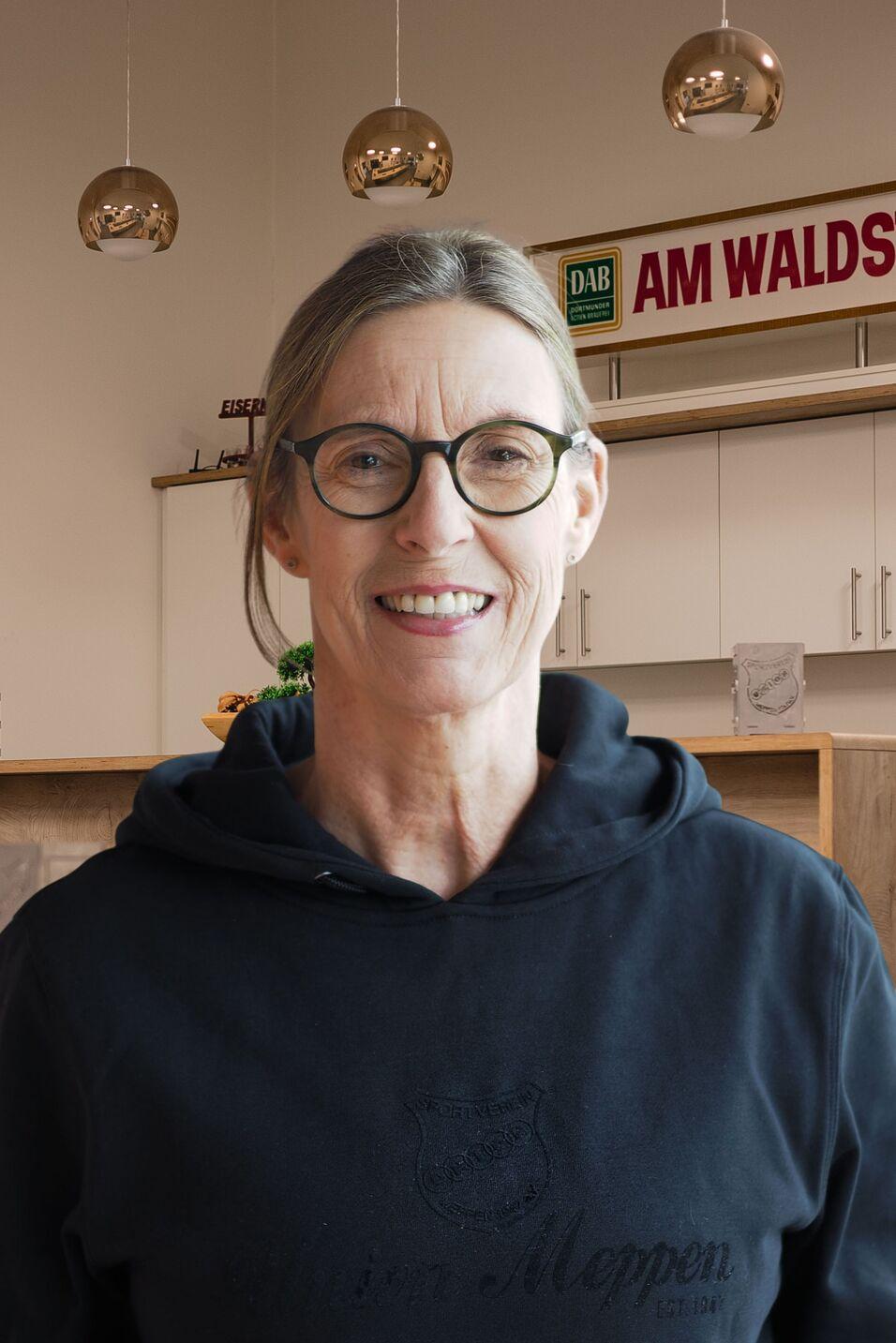 L&auml;chelnde Frau mit Brille in schwarzem Hoodie vor modernem K&uuml;cheninterieur mit Holzregalen und dekorativem Hintergrund.