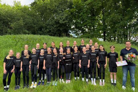 Gruppe von Jugendlichen in schwarzen T-Shirts steht auf einer Wiese, lächelnd mit Blumenstrauß und Plakat in der Mitte.