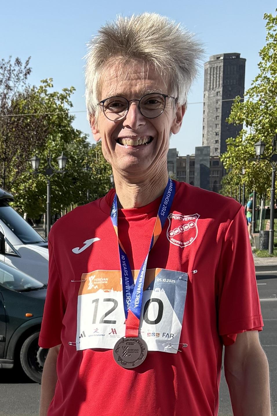 Teilnehmer des Yerevan Marathons 2025 steht mit Medaille vor einem Banner mit Kurs- und Veranstaltungsinformationen.