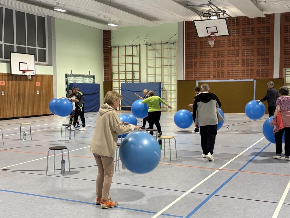 Eine Gruppe von Menschen trainiert in einer Sporthalle mit blauen Gymnastikb&auml;llen und Hockern.