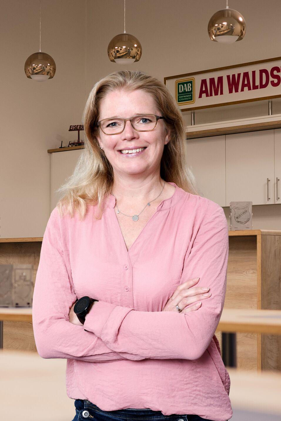 Frau mit blonden Haaren und Brille steht l&auml;chelnd mit verschr&auml;nkten Armen in einem modernen Raum.