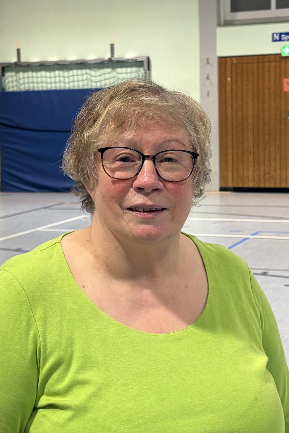 Frau mit Brille und blonden Haaren tr&auml;gt ein gr&uuml;nes Oberteil in einer Sporthalle mit Basketballkorb im Hintergrund.