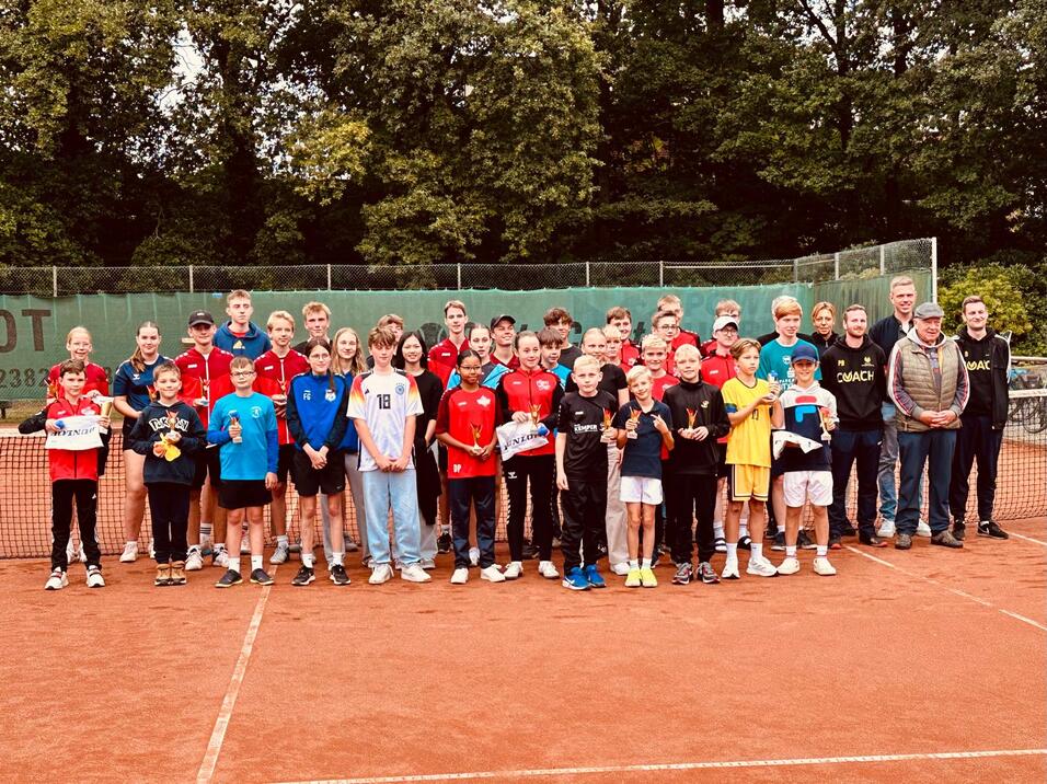 Gruppe von Kindern und Jugendlichen in Sportbekleidung auf einem Tennisplatz, l&auml;chelnd und posierend f&uuml;r ein Foto.