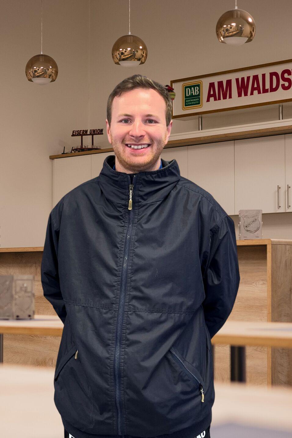 L&auml;chelnder Mann in einem blauen Jacket steht in einem modernen Raum mit Holz- und Metallelementen im Hintergrund.