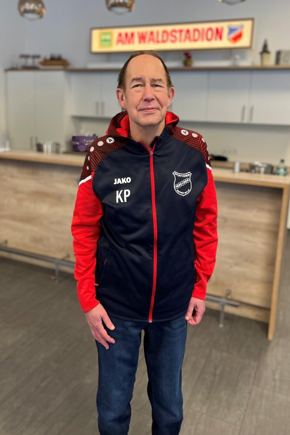 Mann in Sportjacke steht vor einer Theke mit einem Schild &bdquo;AM WALDSTADION&ldquo; im Hintergrund.