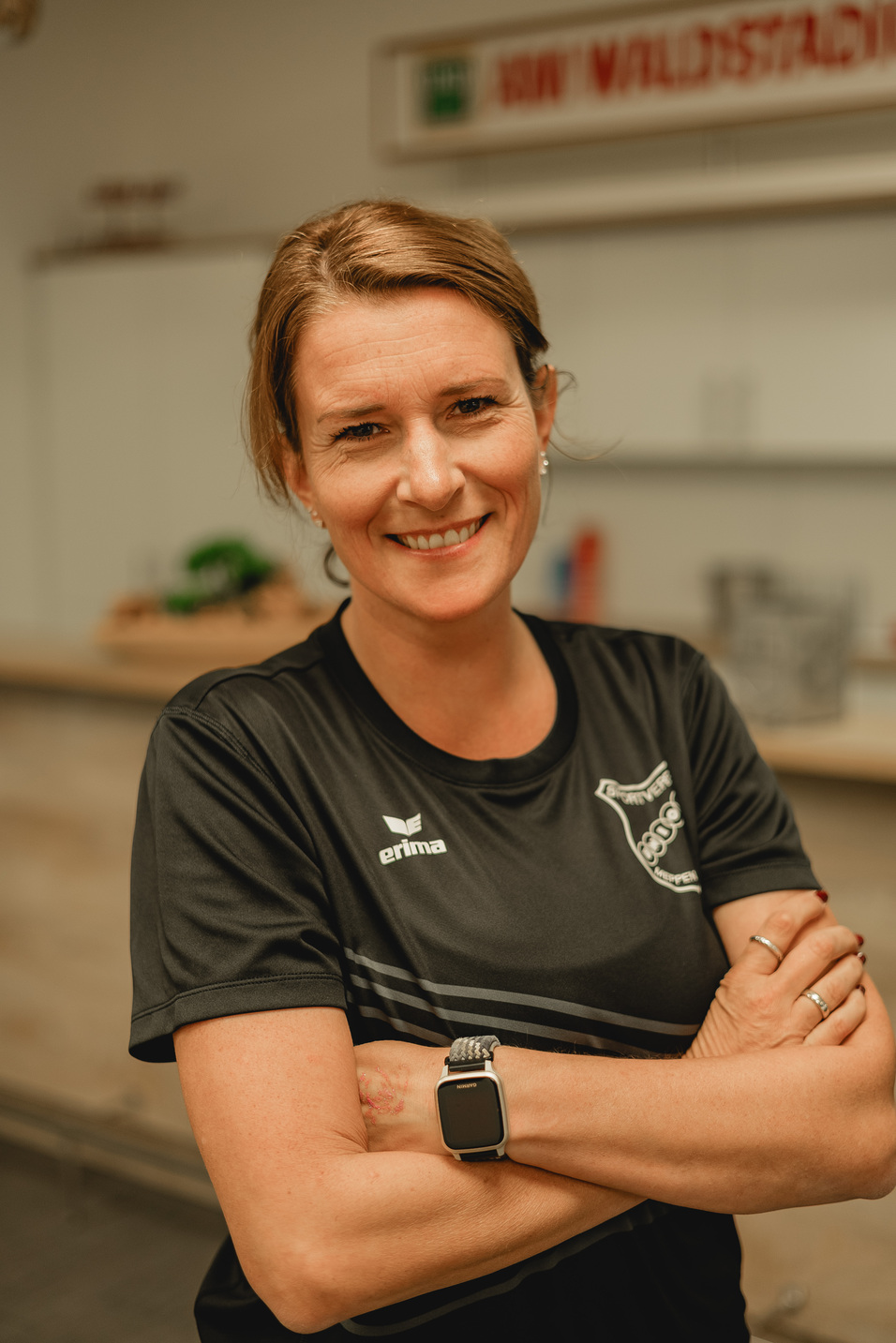 Frau mit kurzem, braunem Haar l&auml;chelt in Sportoberbekleidung und steht mit verschr&auml;nkten Armen in einem offenen Raum.