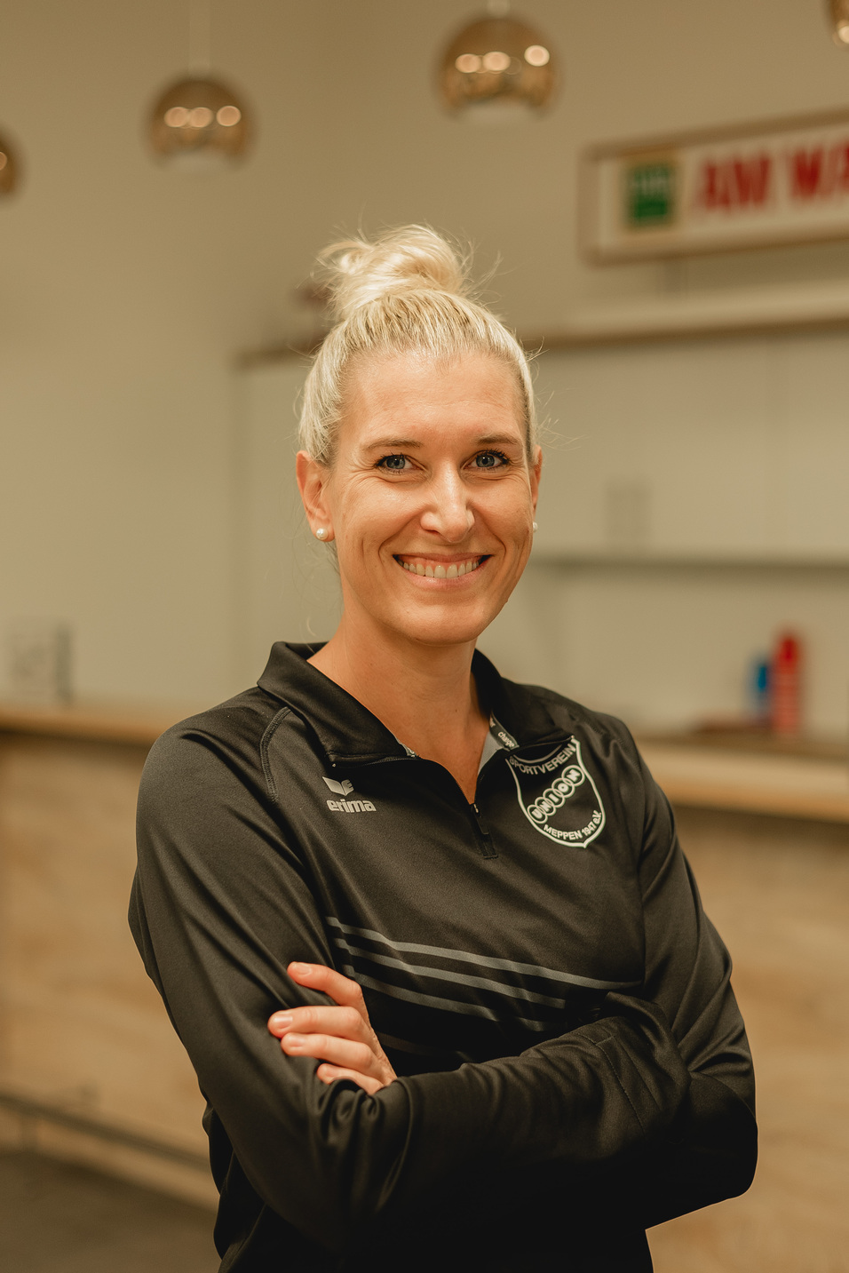 L&auml;chelnde Frau mit blonden Haaren, gekleidet in einem schwarzen Sportoberteil, steht vor einem hellen Hintergrund.