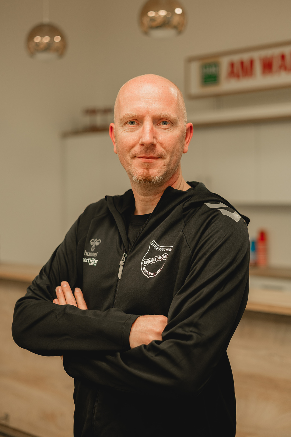 Portr&auml;t eines Mannes in Schwarz mit verschr&auml;nkten Armen, stehend in einer modernen Innenk&uuml;che.