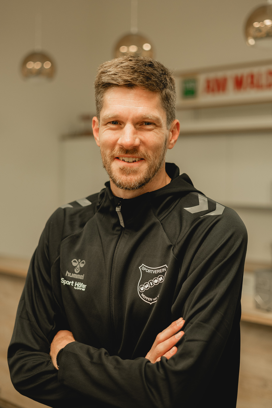 L&auml;chelnder Mann in Sportjacke mit offenem Rei&szlig;verschluss, steht vor einem hellen Hintergrund mit dekorativen Lampen.