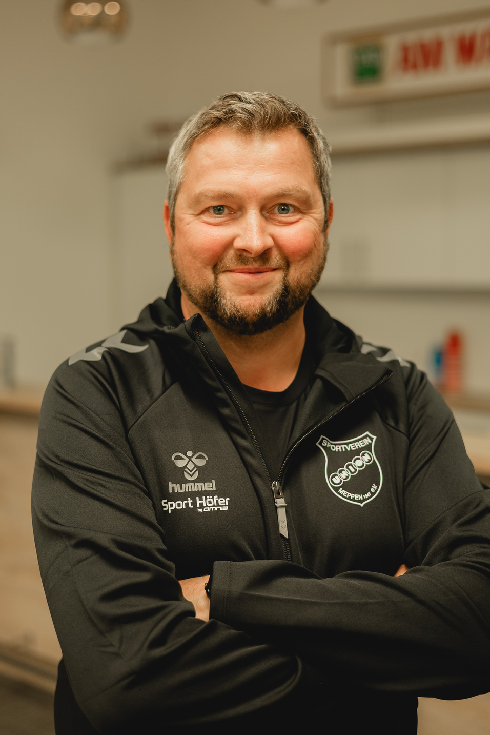 Mann mit kurzem grauem Haar und Bart in schwarzer Sportjacke posiert l&auml;chelnd vor einem hellen Hintergrund.