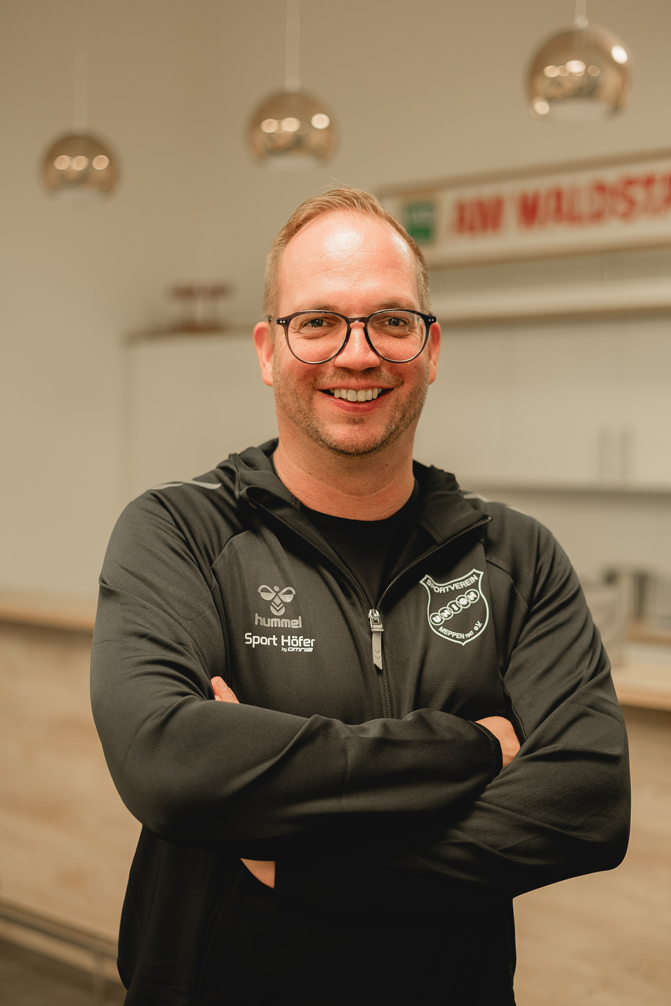 L&auml;chelnder Mann in schwarzer Sportjacke steht mit verschr&auml;nkten Armen in einer modernen Innenraumumgebung.