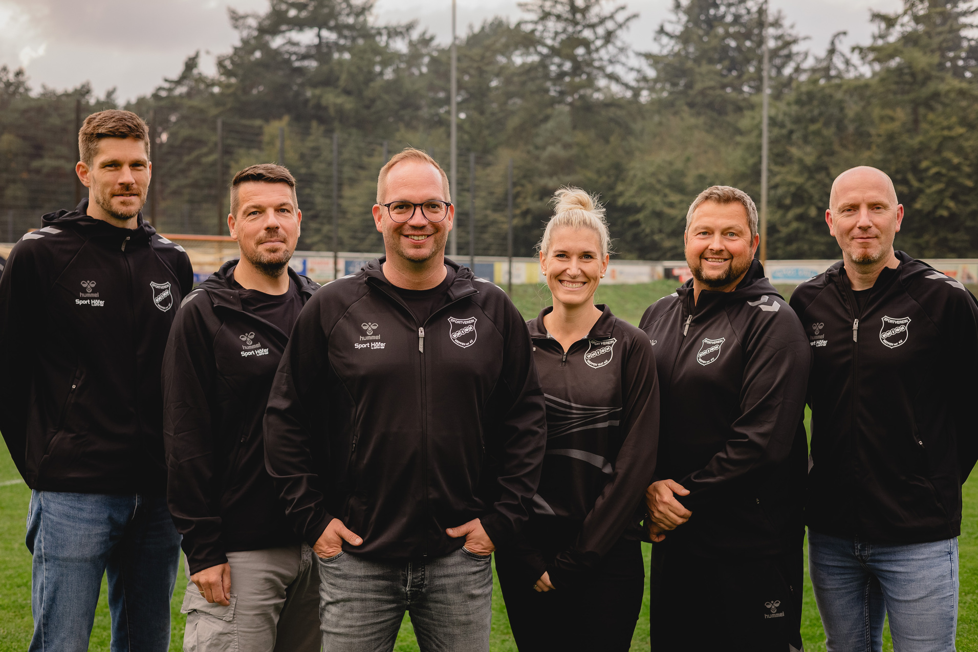 Sechs Personen in Sportbekleidung stehen auf einem Fu&szlig;ballfeld, umgeben von B&auml;umen und einem sportlichen Ambiente.