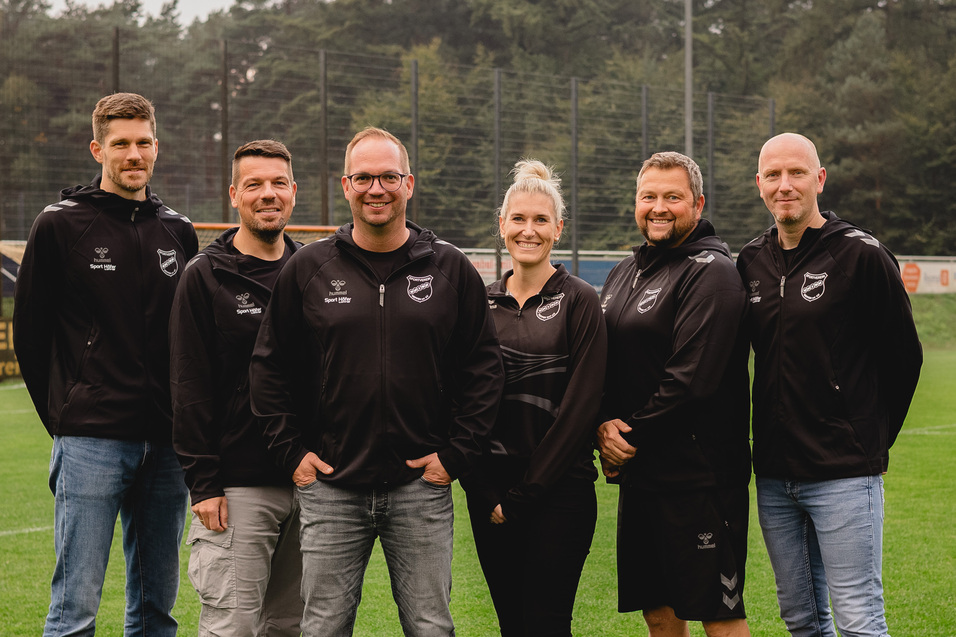 Gruppenfoto von sechs Personen in Sportbekleidung auf einem Fu&szlig;ballfeld, mit B&auml;umen im Hintergrund.