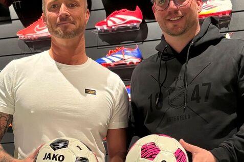 Zwei Männer stehen in einem Sportgeschäft und halten jeweils einen Jako-Fußball in der Hand. Im Hintergrund Schuhe.
