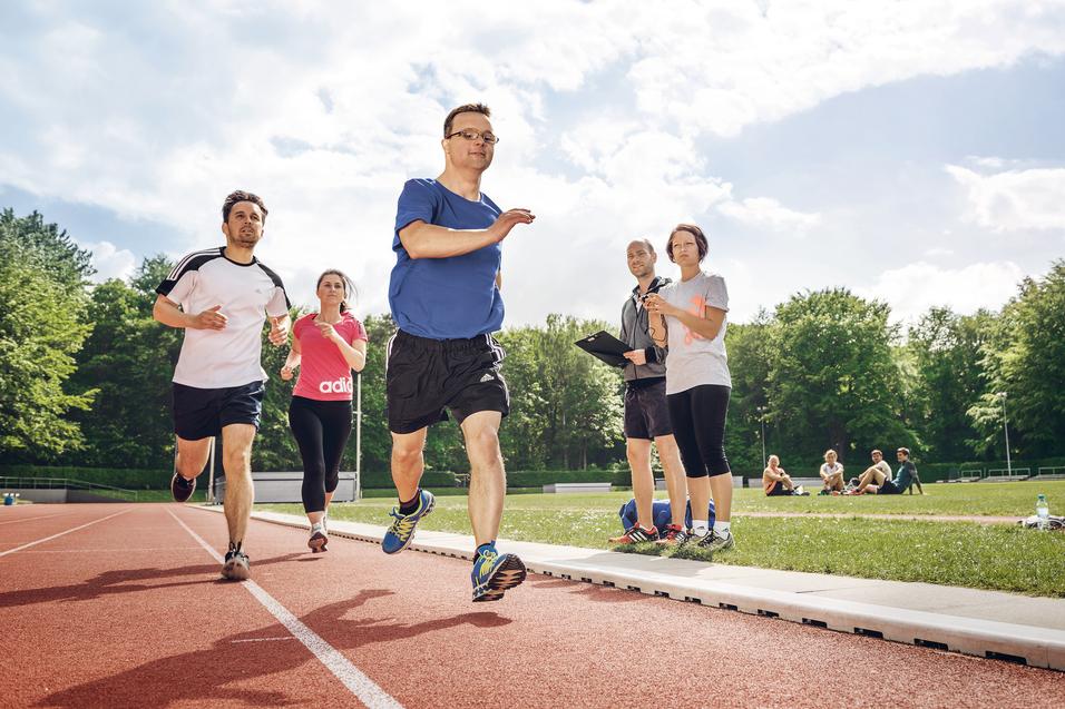 Inklusive Laufgruppe mit Läufer mit Prothese auf der Bahn; zwei Personen beobachten mit Stoppuhr und Klemmbrett.