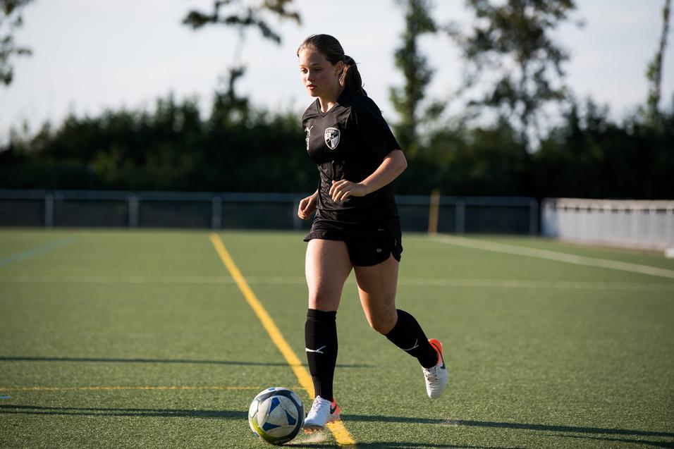 Eine Frau in schwarzer Sportkleidung dribbelt einen Fußball auf einem Kunstrasenplatz.