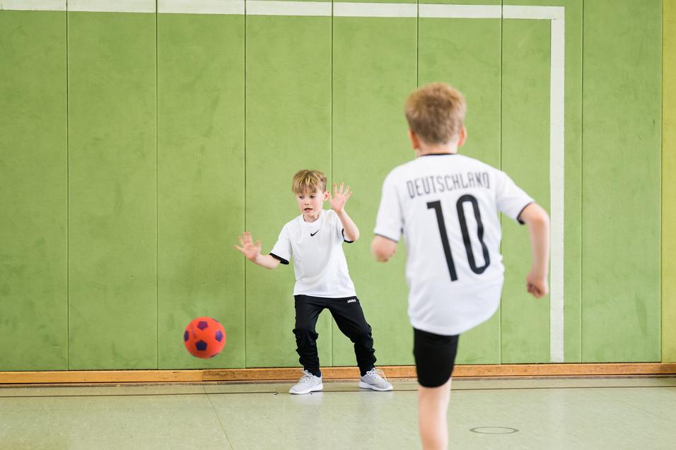 Zwei Kinder spielen in einer Turnhalle mit einem leichten, roten Ball, während eine grüne Wand im Hintergrund sichtbar ist.