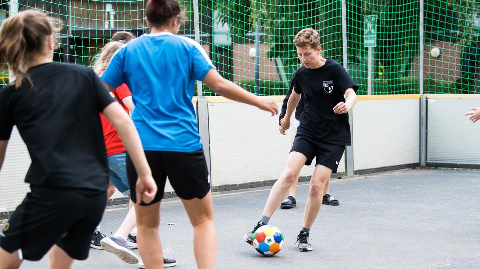 Gruppe von Jugendlichen spielt Fußball in einem umschlossenen Sportbereich mit Netz und Gummiuntergrund.