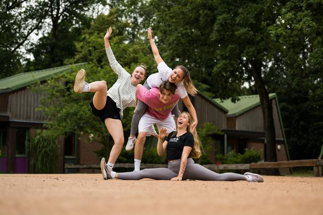 Vier junge Frauen posieren fröhlich auf einer sandigen Fläche, eine sitzt im Spagat, während die anderen sie umarmen.