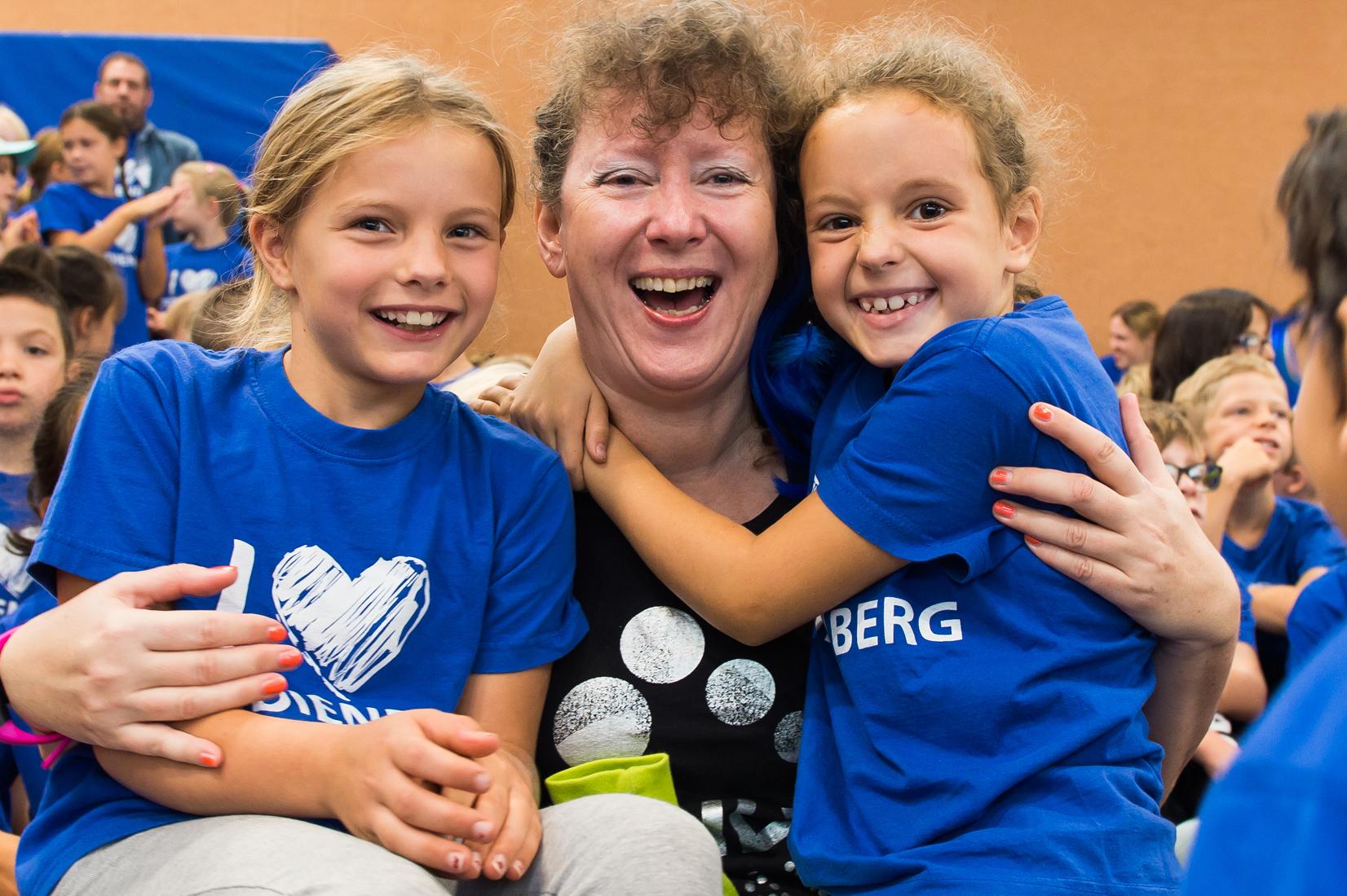 Lächelnde Kinder umarmen eine Frau in einem schwarzen Shirt, alle tragen blaue T-Shirts in einer Gruppenaktivität.