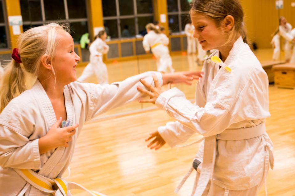 Zwei Mädchen im Karateanzug üben spielerisch im Trainingsraum miteinander, umgeben von weiteren Trainierenden.