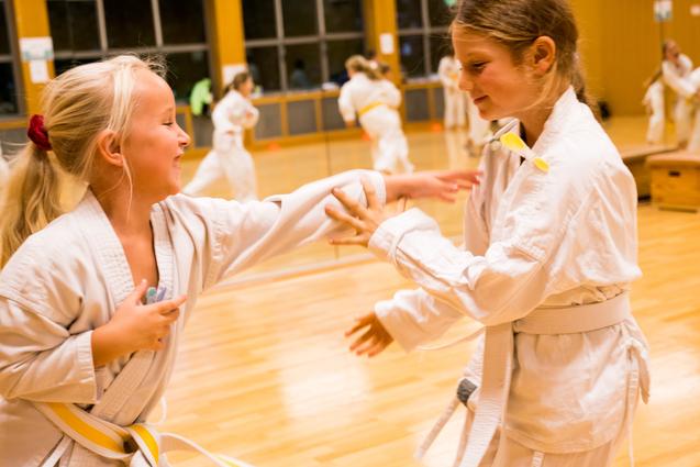 Zwei Mädchen im Karateanzug üben spielerisch im Trainingsraum miteinander, umgeben von weiteren Trainierenden.