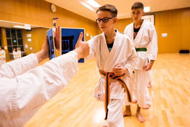 Junge in Karate-Gi mit orangenem Gürtel im Training, empfängt Trainingsmaterial von einem Trainer. Hintergrund mit weiteren Schülern.