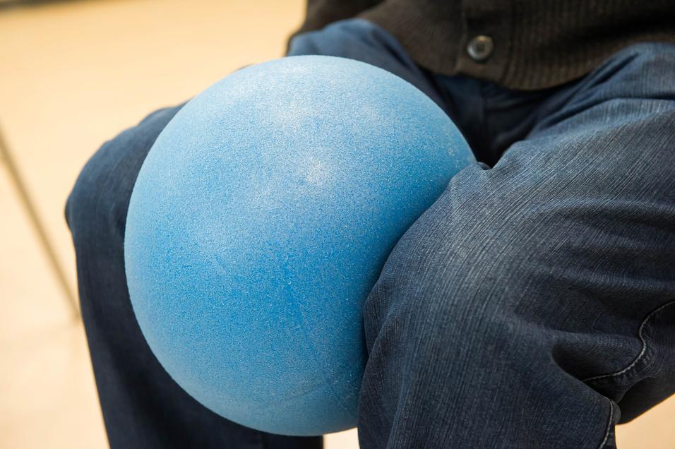 Blauer Gymnastikball ruht auf den Knien einer Person in blauer Jeans. Unschärfe im Hintergrund.