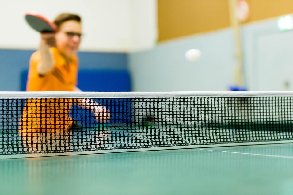 Tischtennisschläger in der Hand eines Spielers, im Hintergrund ein Ball über das Netz fliegend auf einer grünen Tischtennisplatte.