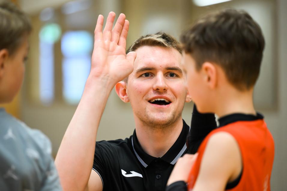 Trainer spricht mit einem jungen Spieler, während er seine Hand hebt, in einer Sporthalle mit Holzwand.