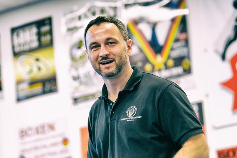 Mann mit Boxhandschuhen in einem Trainingsraum, umgeben von Sportplakaten und einem Boxring.