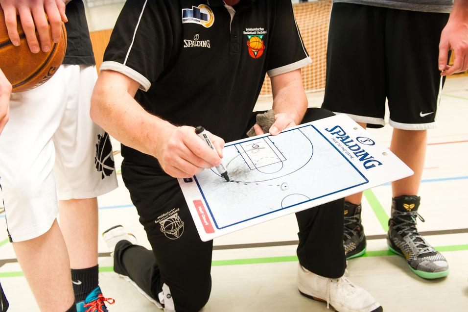 Trainer kniet mit Taktiktafel und zeichnet Spielzüge, während Spieler im Hintergrund stehen und einen Basketball halten.