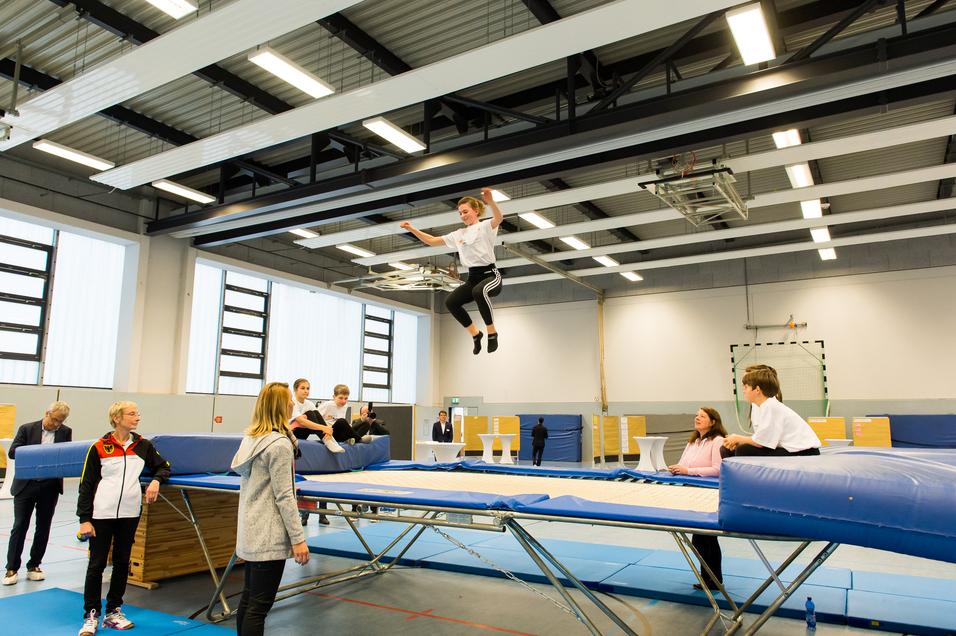Junge springt auf einem Trampolin, während Zuschauer und Trainer im Hintergrund beobachten. Sporthalle mit hellem Licht.