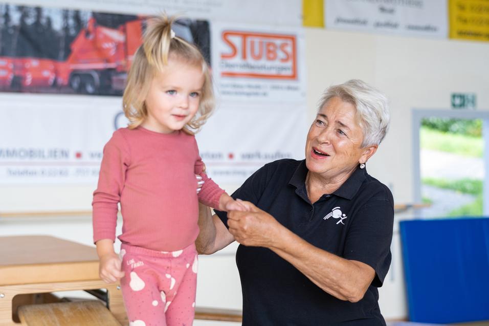 Ein kleines Mädchen steht auf einem Turngerät und wird von einer lächelnden älteren Frau unterstützt.